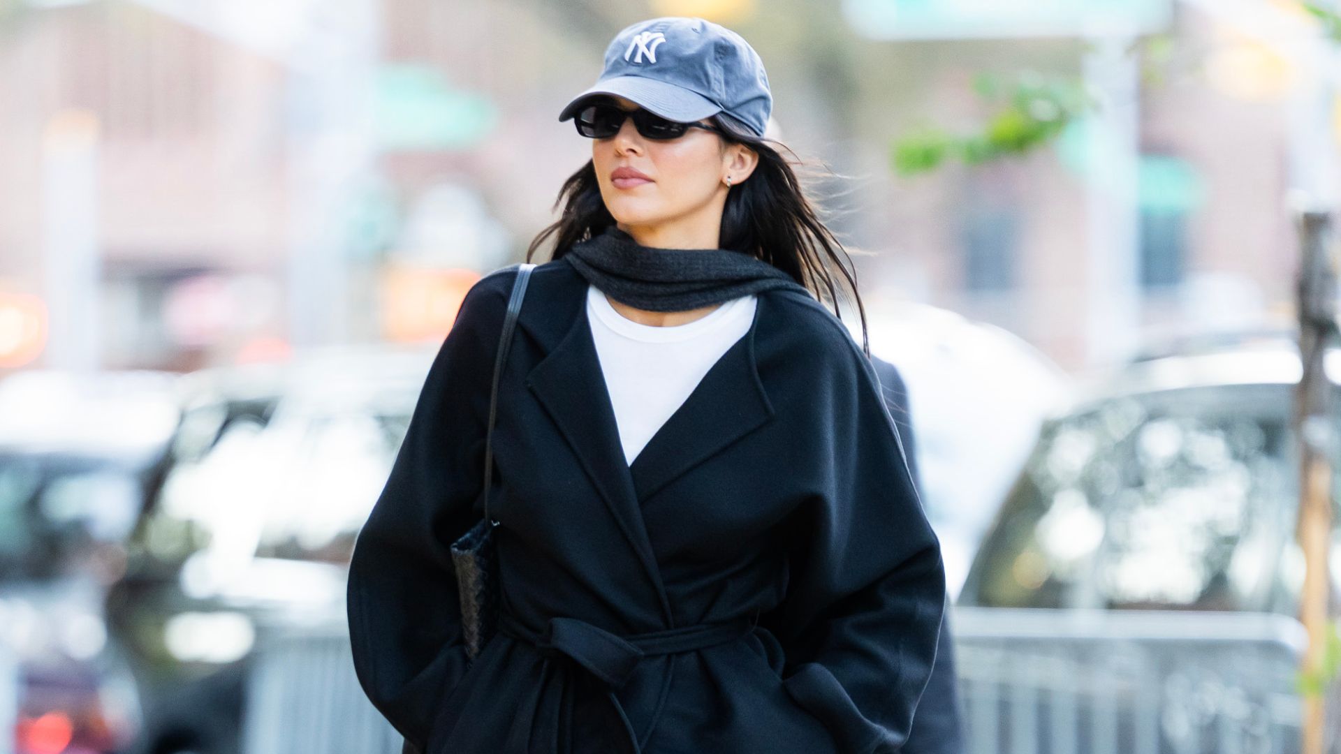 Kendall Jenner walks down the street wearing bootcut jeans with pointed-toe boots and a black belted coat, grey scarf and cap.
