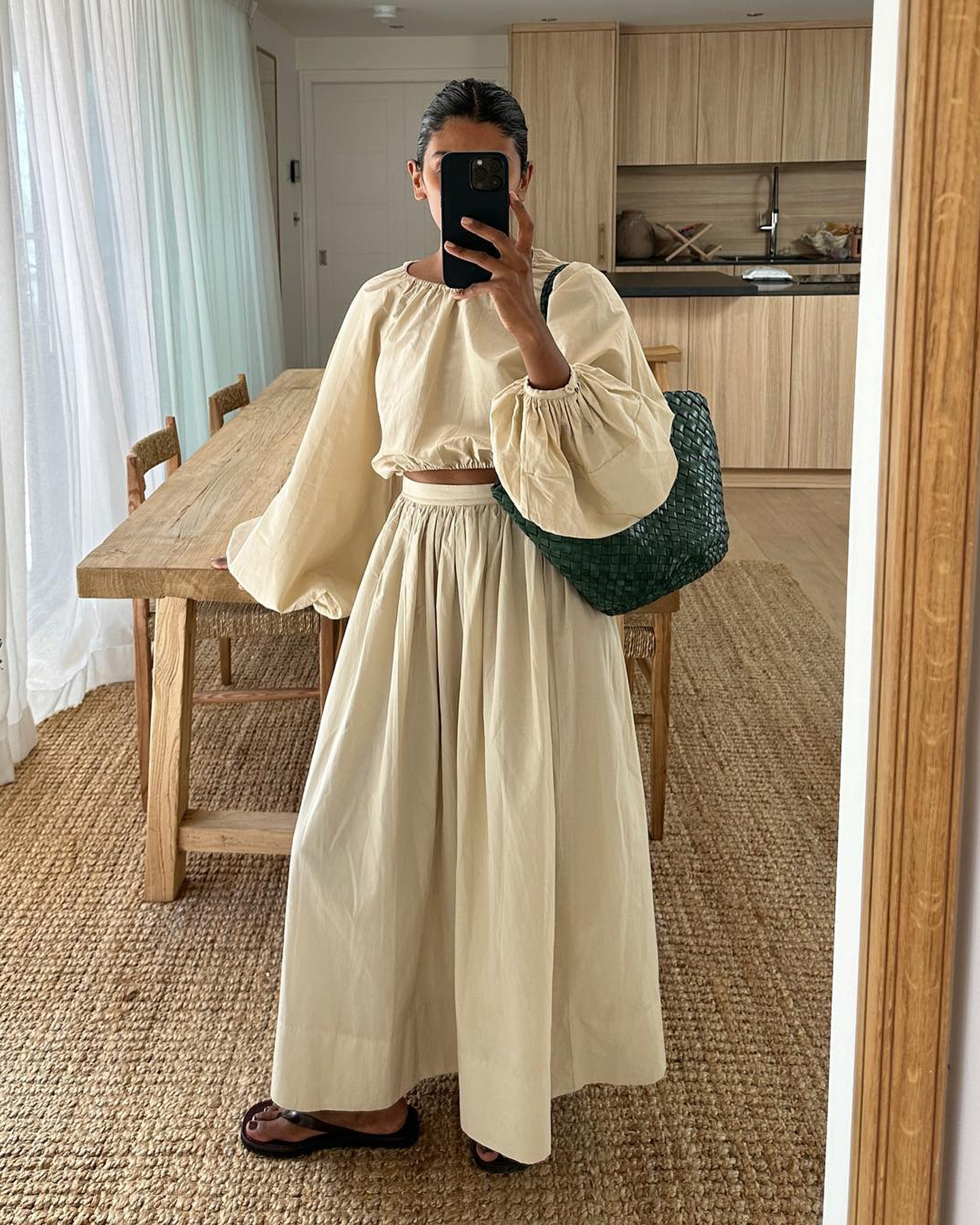 British style influencer Monikh Dale poses for a mirror selfie in the living room of her London home wearing a beige linen smocked neck cropped top with balloon sleeves and a matching linen skirt, green Dragon Diffusion tote bag, and black flat flip-flop sandals.