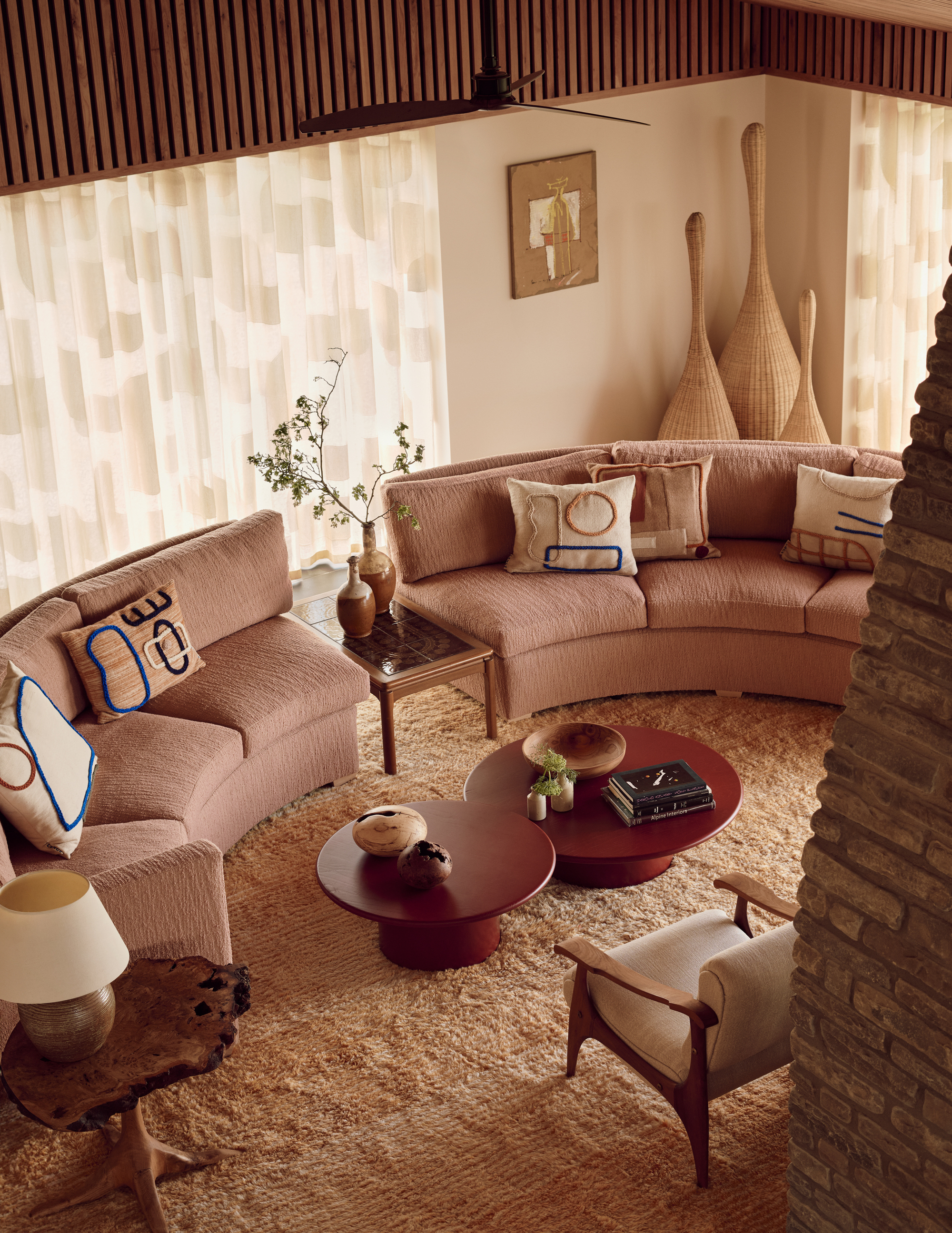 Image of a living room from above with a blush pink curved sofa and a beige shag rug. There are sheer curtains on the back window and in front of the sofa is a red circular nesting coffee table.