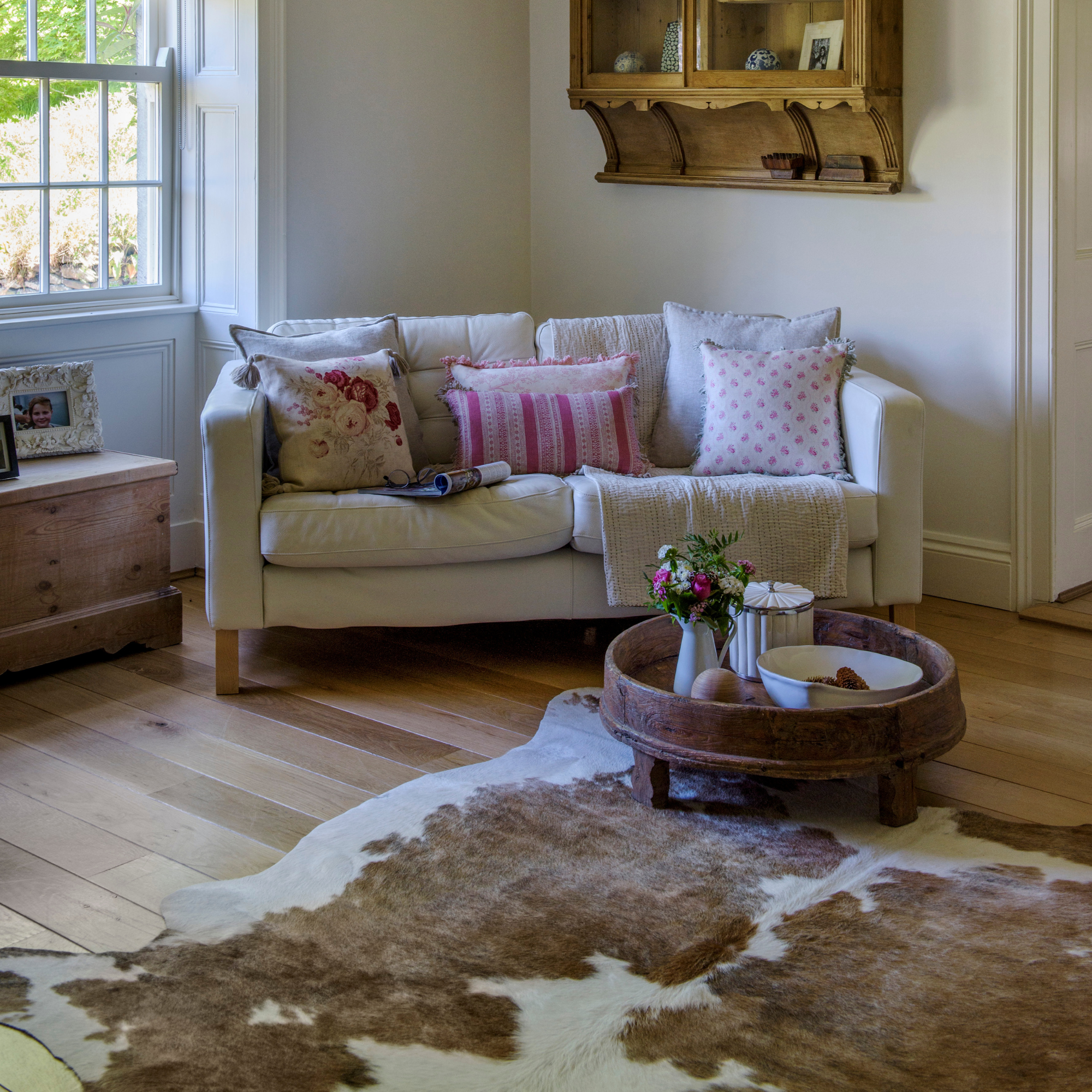 A white living room with sofa and antique rustic style table and animal hide rug on a wooden floor.