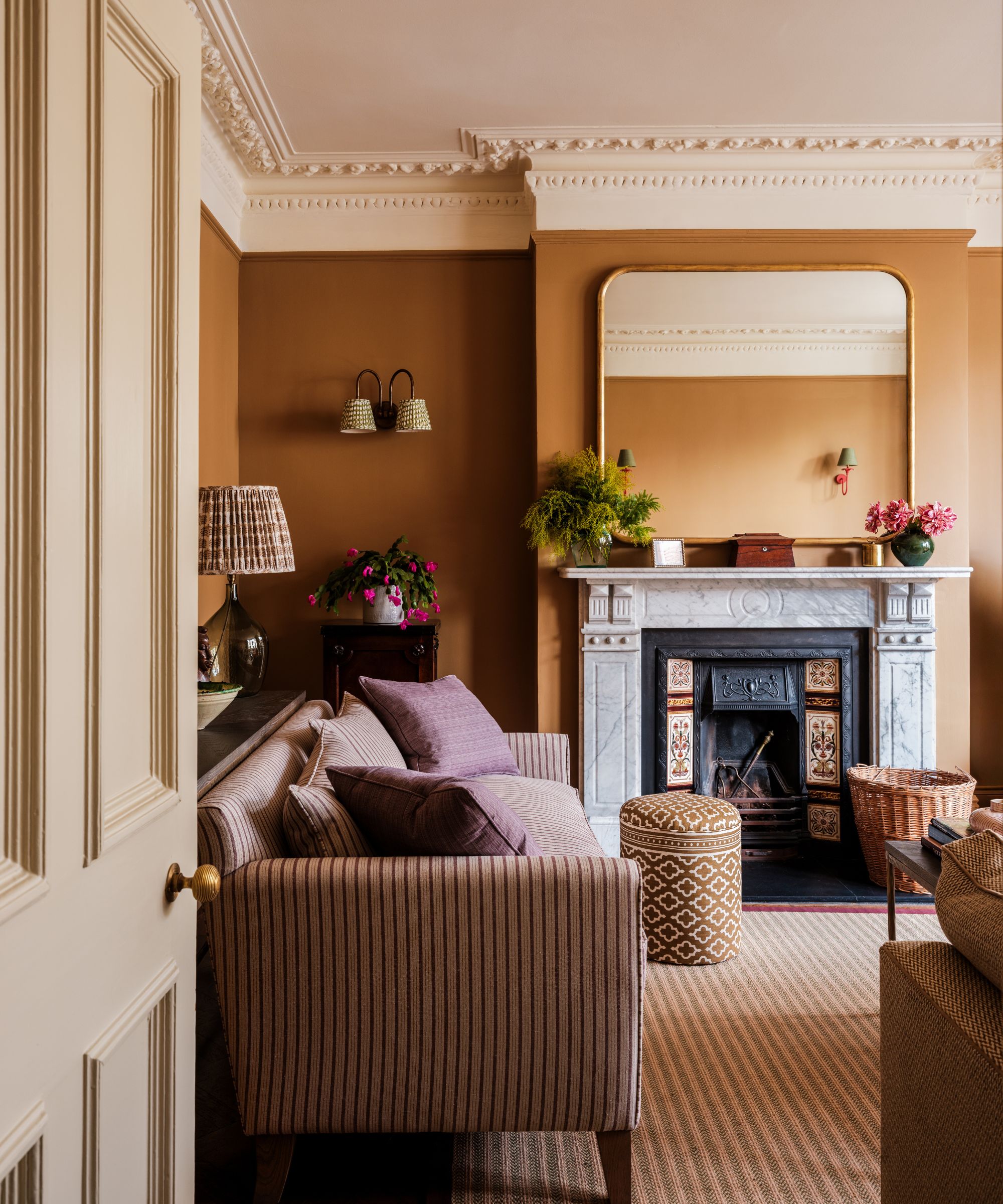 A traditional living room with rich ochre walls, a white ceiling with moldings, and a large marble fireplace with a mirror above. A striped rug, a striped sofa, and warm patterned decor.