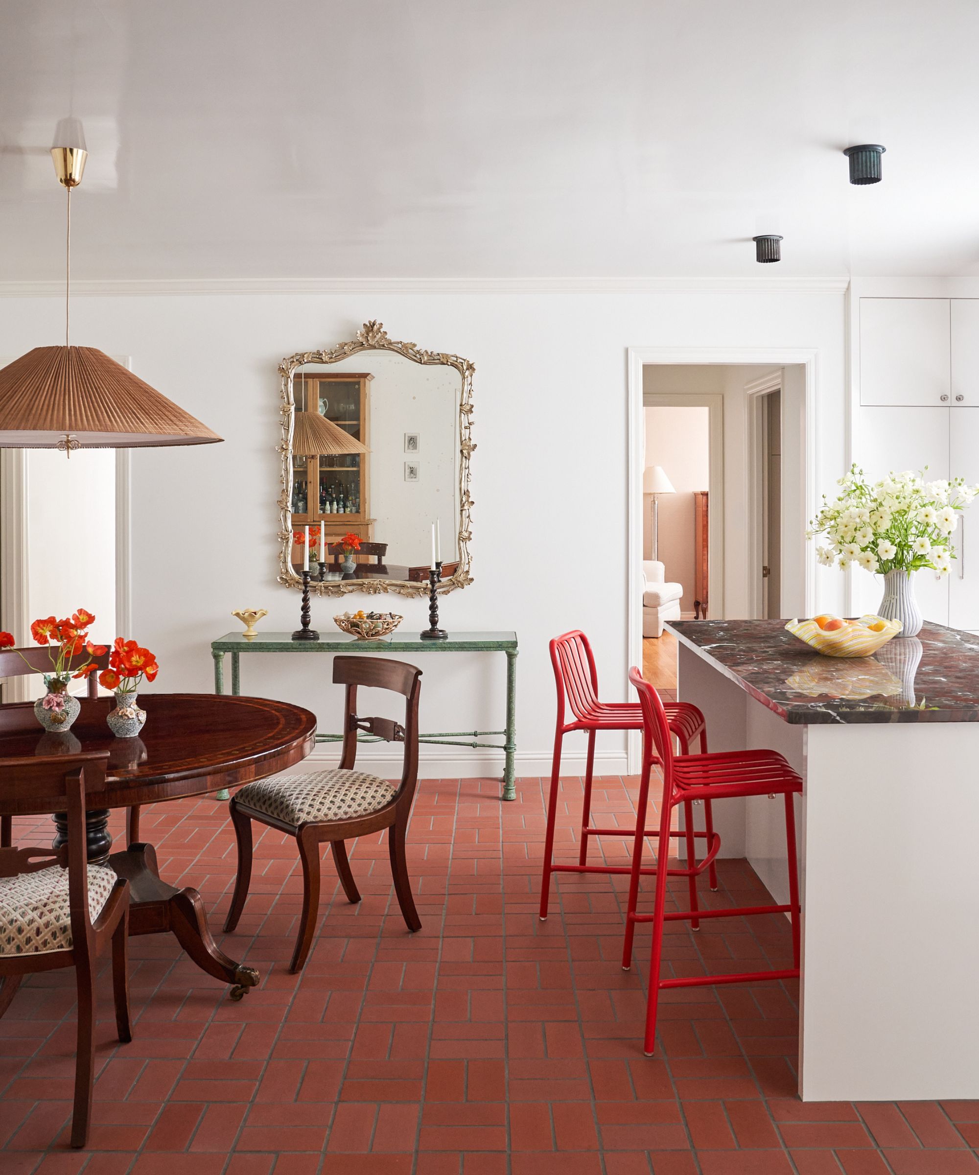 a white painted kitchen with white cabinetry, brick tiled floor, dark red marble island with red stools and an antique wooden round table and chairs
