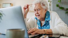 An older woman holds her glasses up as she looks at her laptop.