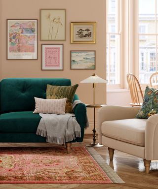 A living room with beige walls with wall art prints on, a dark green velvet couch, a cream armchair, a gold lamp in the middle of them, and a peach patterned rug underneath