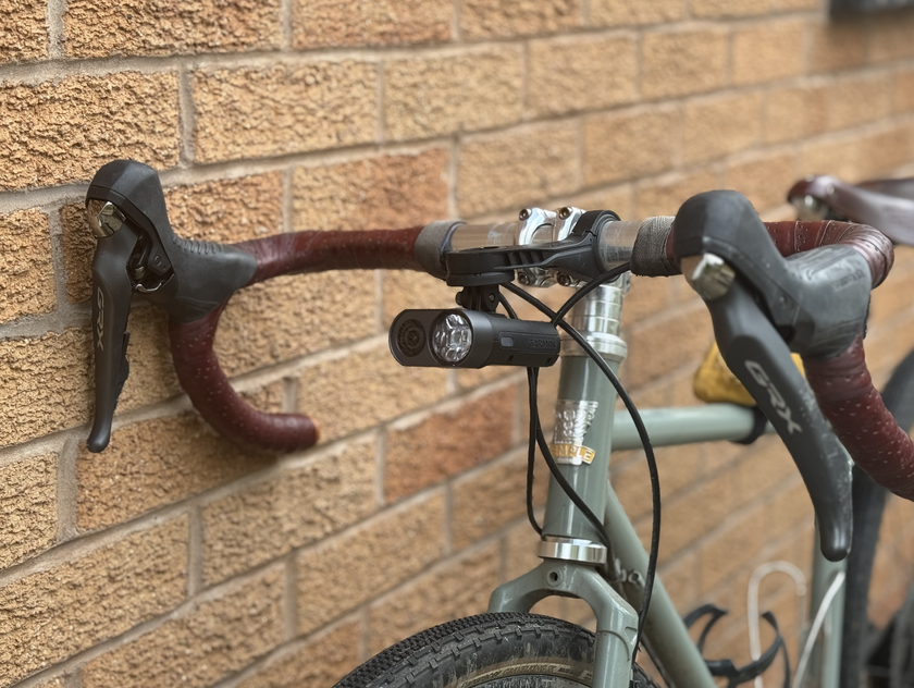 A black bike light attached out front to the handlebars with a small camera on the left and the bike light on the right. It is attached beneath a garmin mount. 