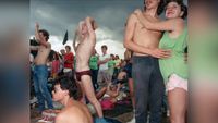 At the 1986 Wackersdorf anti-nuclear protest, a shirtless man raises his arm mid-shout as a couple embrace beside him, a dark storm-clouded sky filling the background.