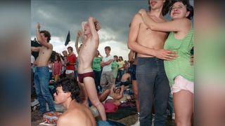 At the 1986 Wackersdorf anti-nuclear protest, a shirtless man raises his arm mid-shout as a couple embrace beside him, a dark storm-clouded sky filling the background.