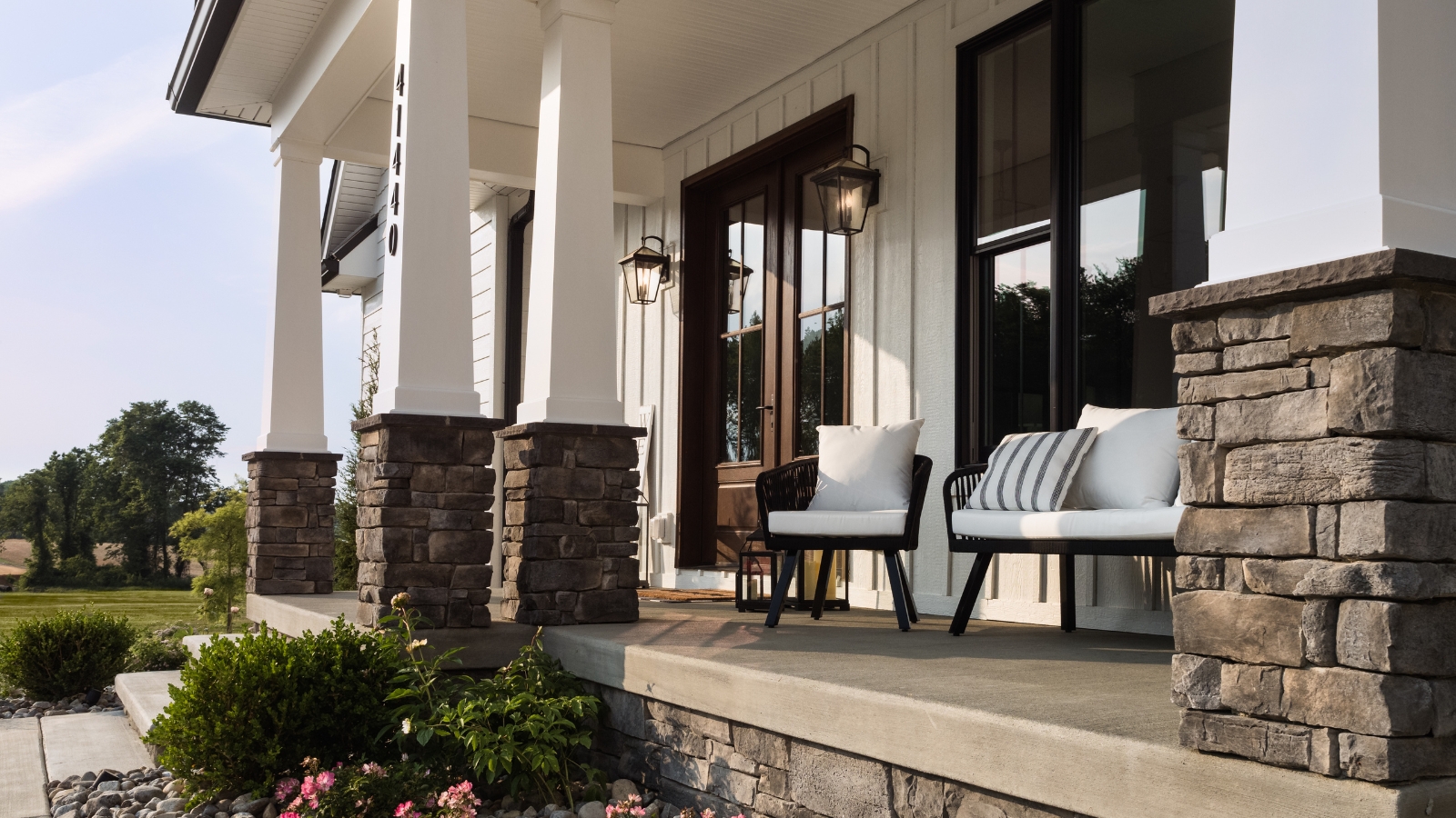 Front porch of stone house with large pillars, two armchairs, and steps leading up to brown double doors.