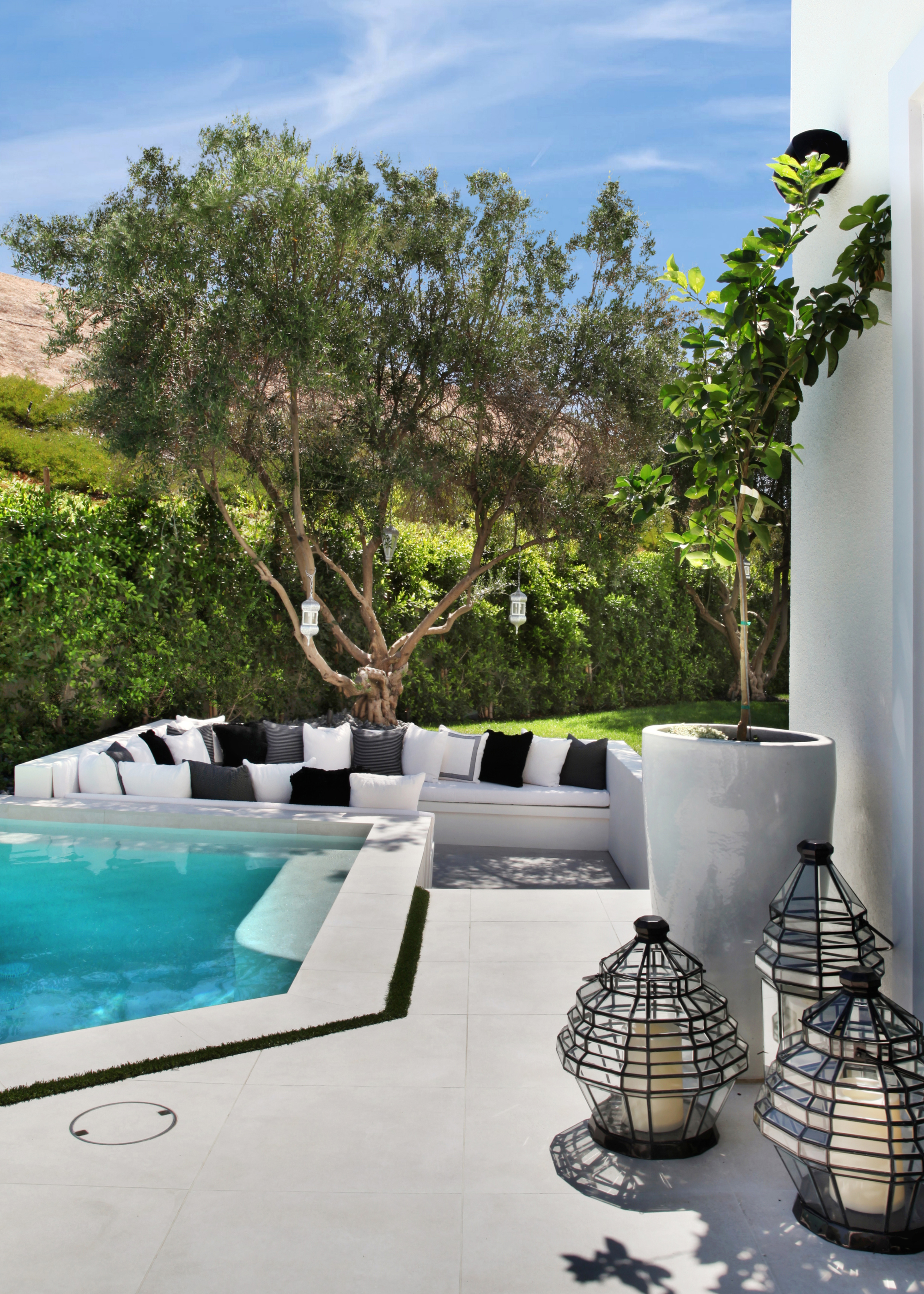 A modern California-style garden with a built-in outdoor seating area with black, white, and gray cushions by the pool with a large tree behind, and three black-wired lanterns with candles are next to the doorway by a potted tree
