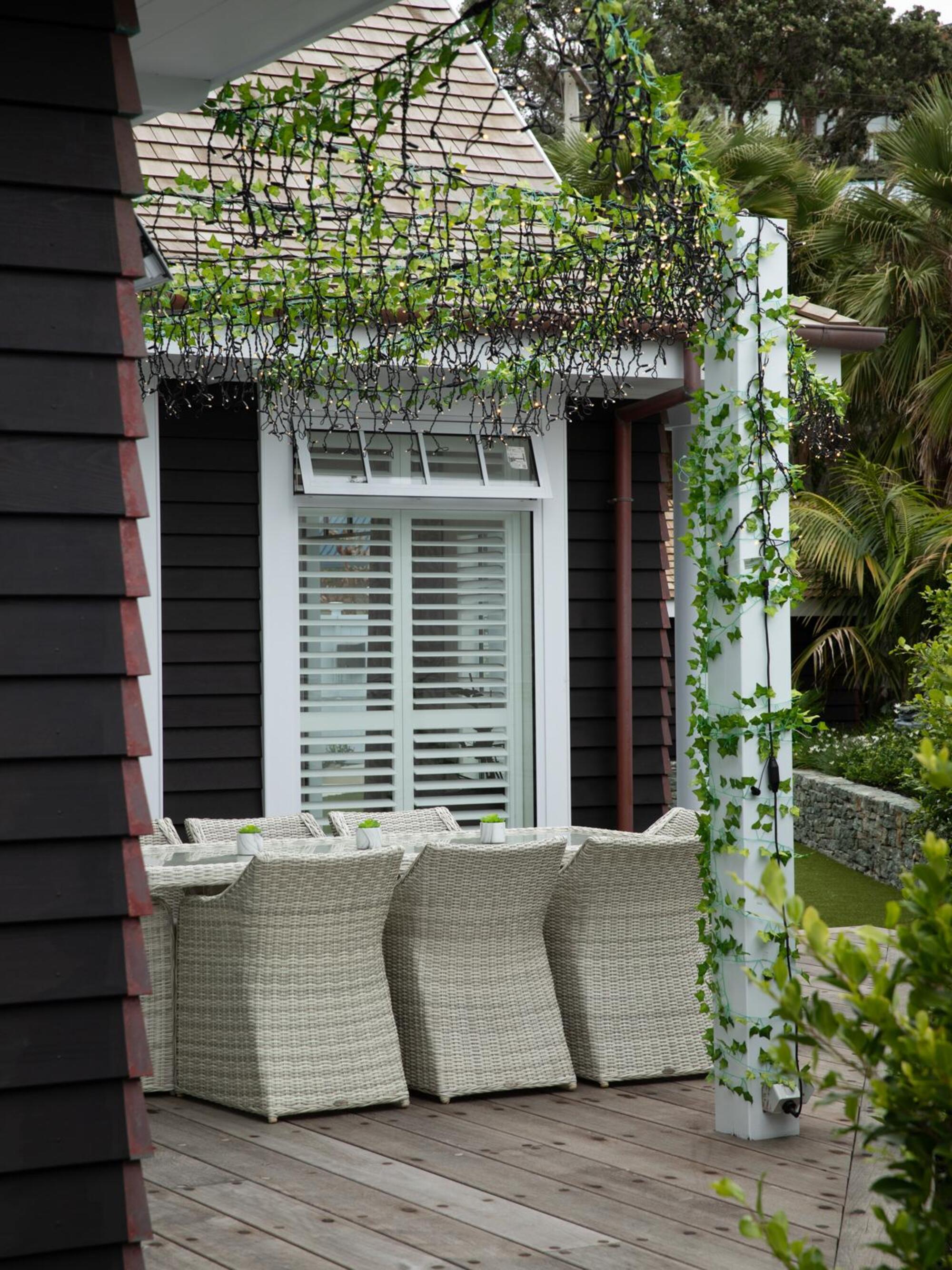small decked outdoor area with pergola with climbing plants and string lights with dining table with cane chairs and black weatherboard home exterior