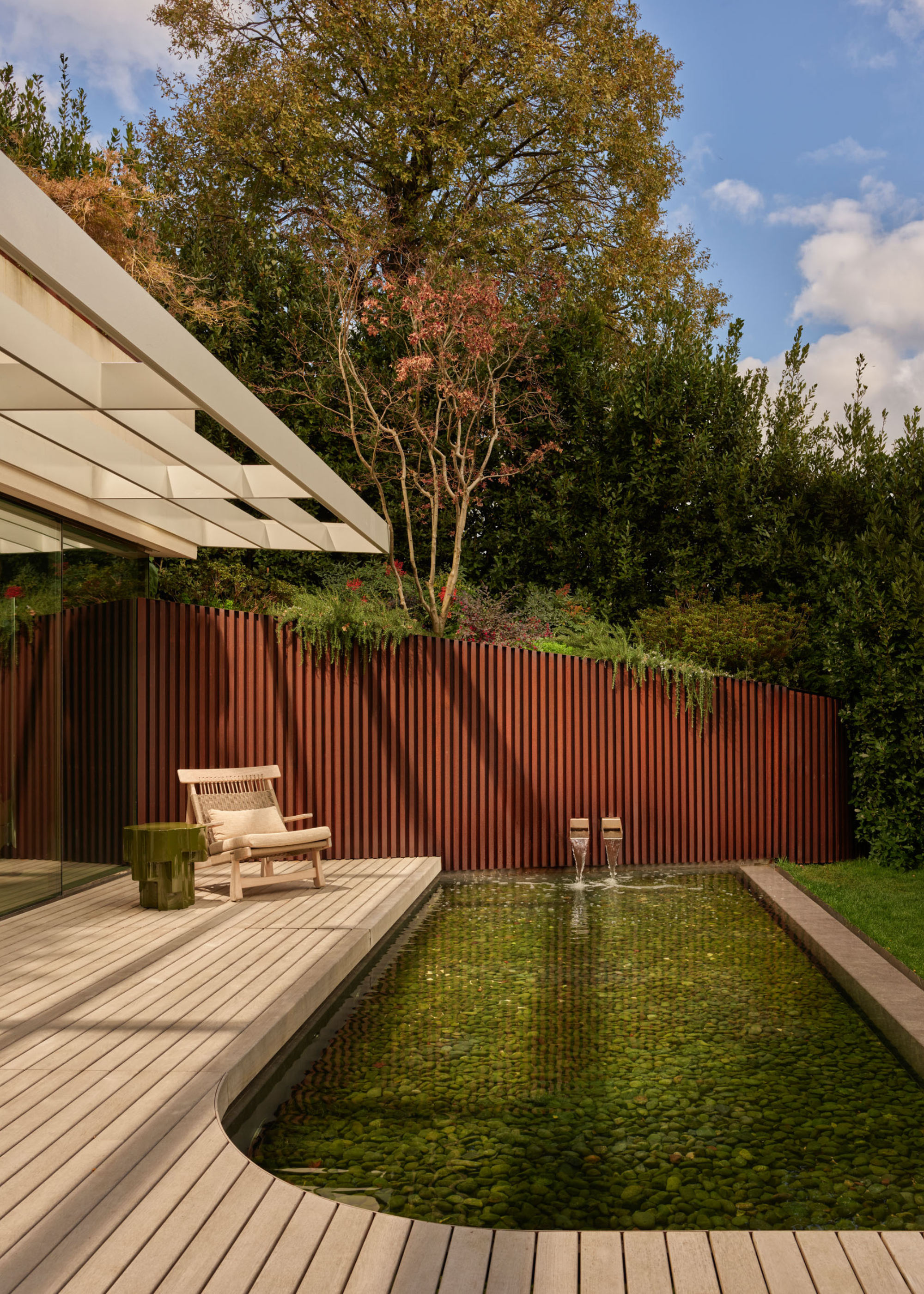 An outdoor space with a pond and a pair of fountain spouts on a wood deck by a lounging outdoor armchair and a side table against a lush backdrop