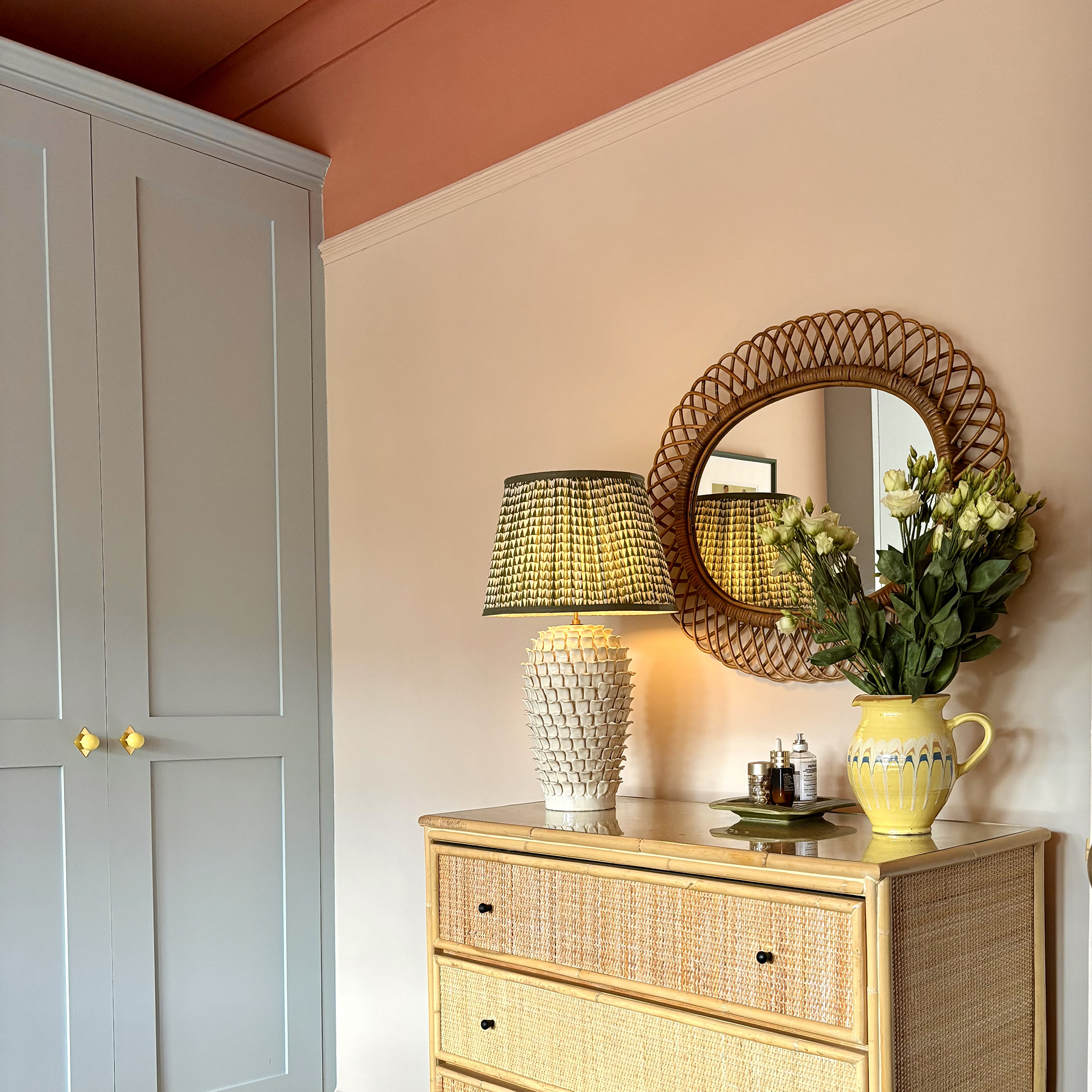 Corner of a bedroom with soft blue painted built-in wardrobes, terracotta ceililng and vintage chest of drawers topped with a vintage lamp and jug