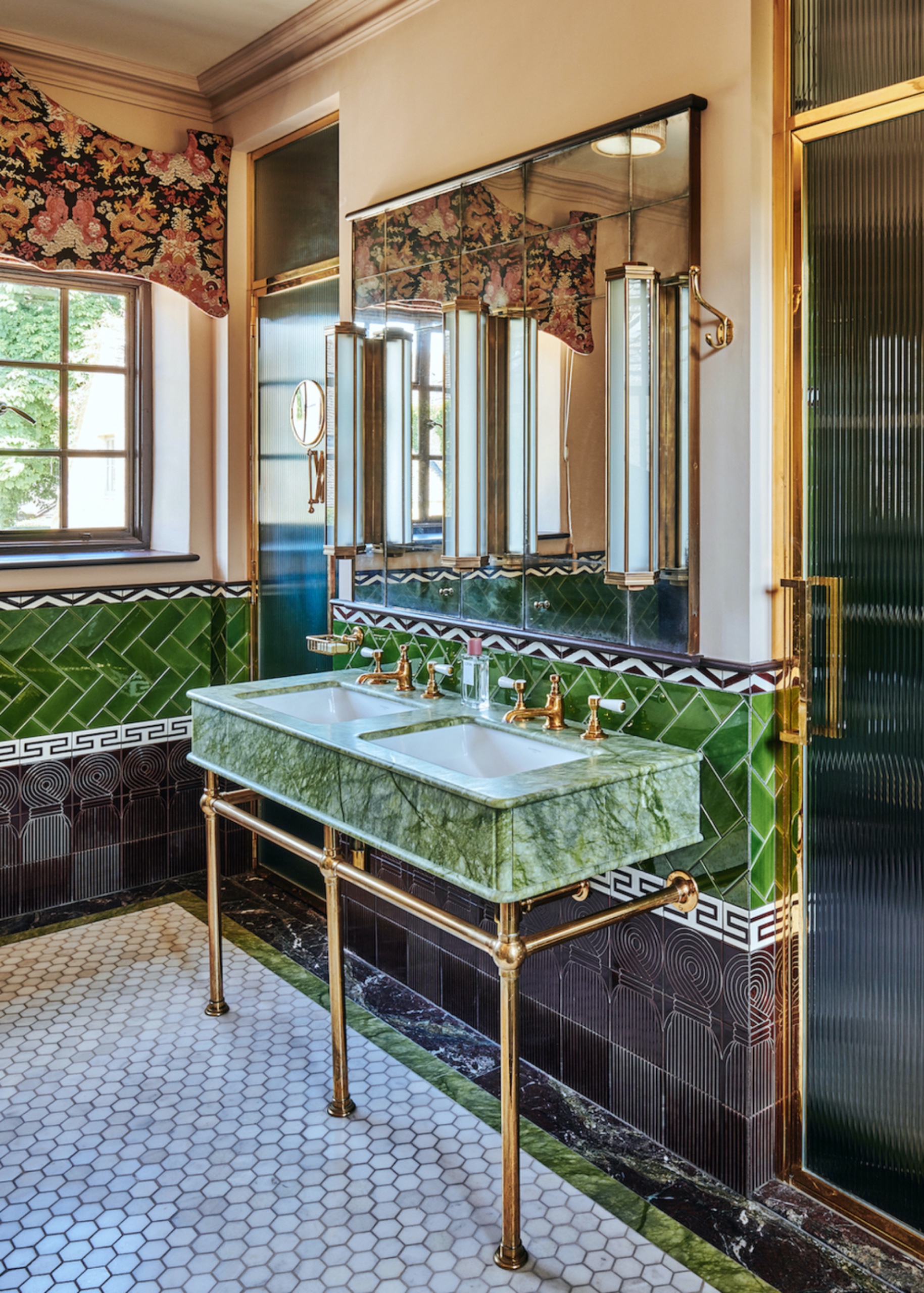 Bathroom with green herringbone wall tiles, decorative borders and dark patterned lower tiles, a green marble double sink, brass fixtures and white hexagonal floor tiles.