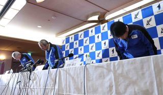 three men in blue tracksuits bow in front of a table lined with microphones