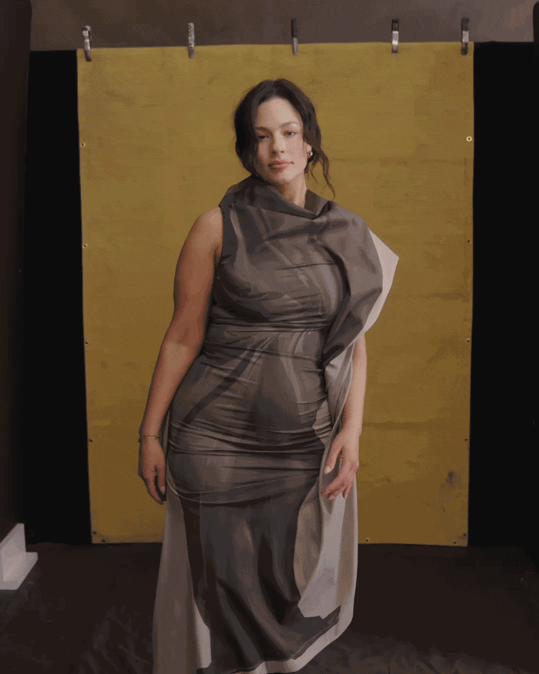 Ashley Graham in a gif wearing a brown couture gown and boots posing on a stool and standing against a yellow backdrop