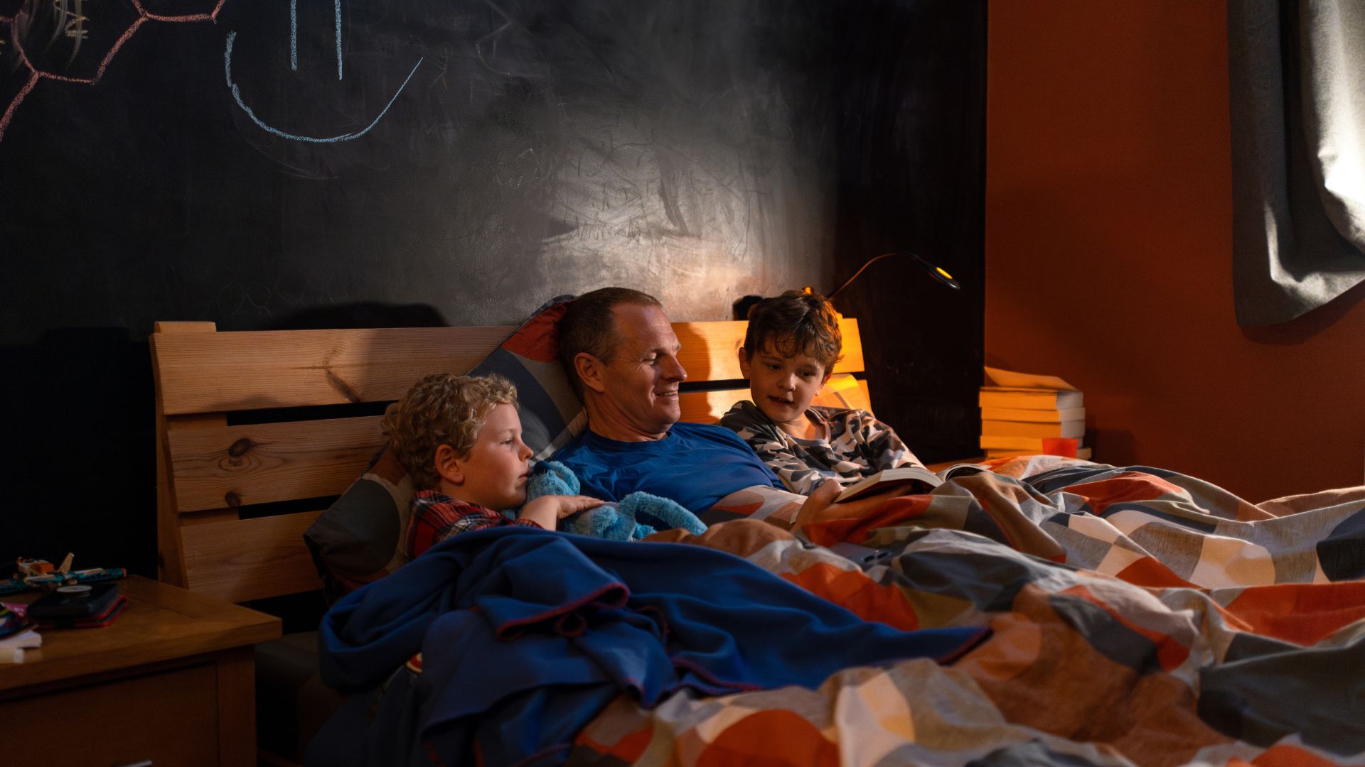 A father reading a book to two children as they all sit in bed under the duvet.