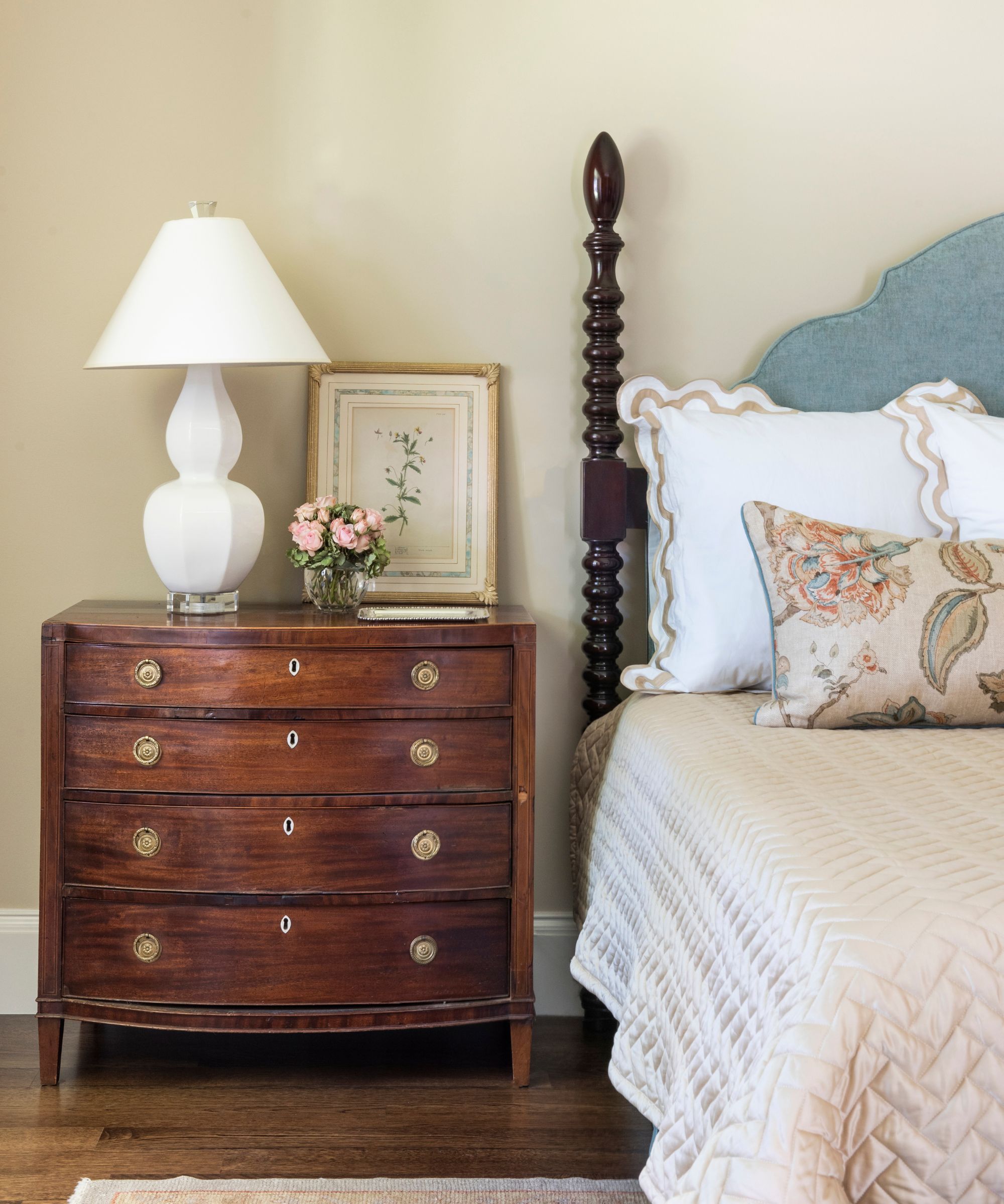 vintage bedroom with soft neutral walls, a wooden turned bed with a blue upholstered headboard, and white scalloped bedding with a quilt and an antique chest bedside table