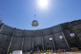 a crane lowers a cylindrical object toward the ground inside a partly finished cement building