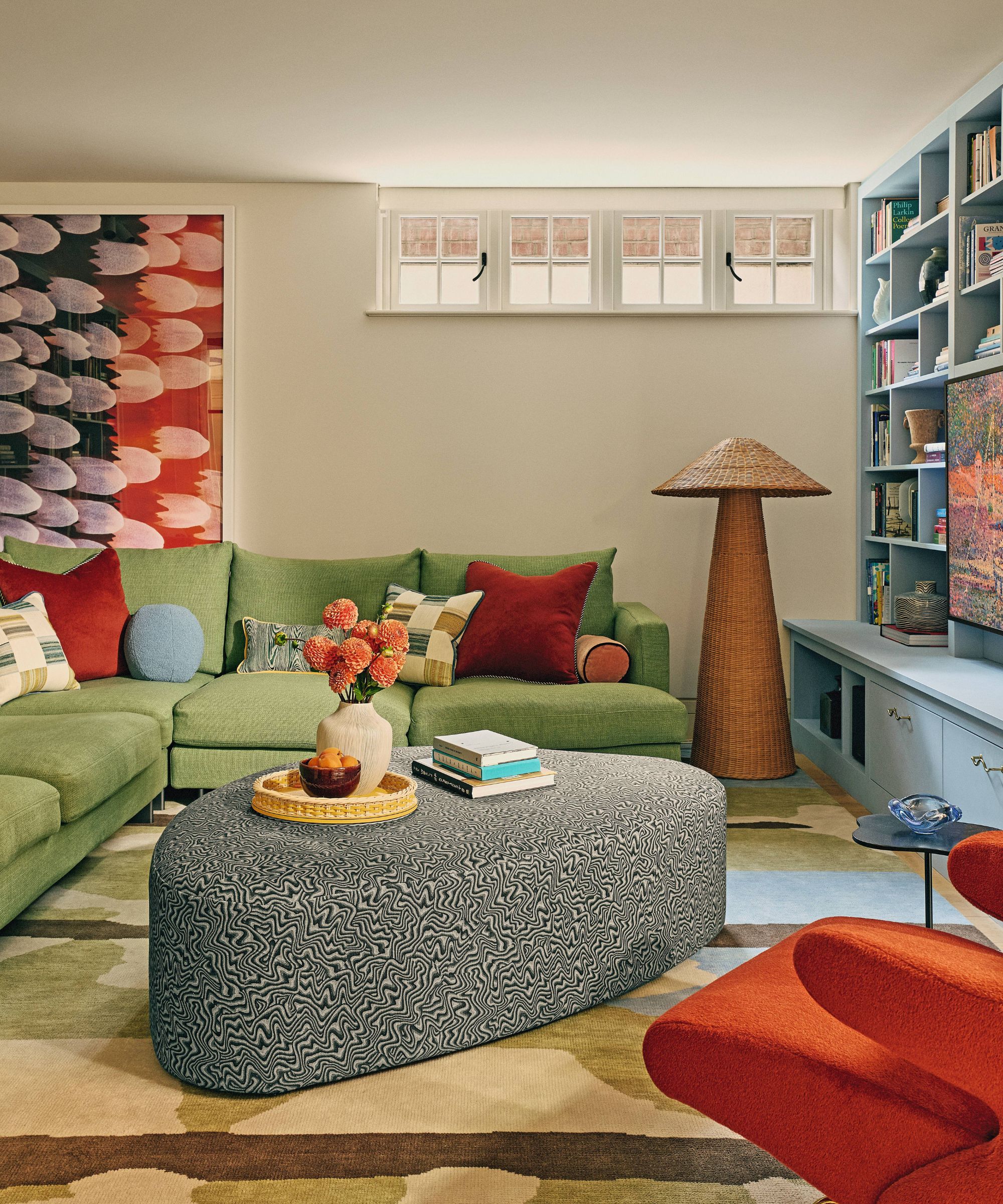 a neutral basement family room with blue media wall cabinetry, a large green sofa, rattan floor lamp, upholstered ottoman and geometric rug