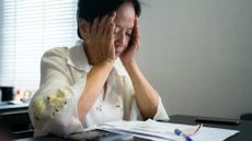 Senior woman sitting at living room table with her head in her hands, calculating expenses