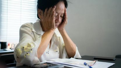 Senior woman sitting at living room table with her head in her hands, calculating expenses