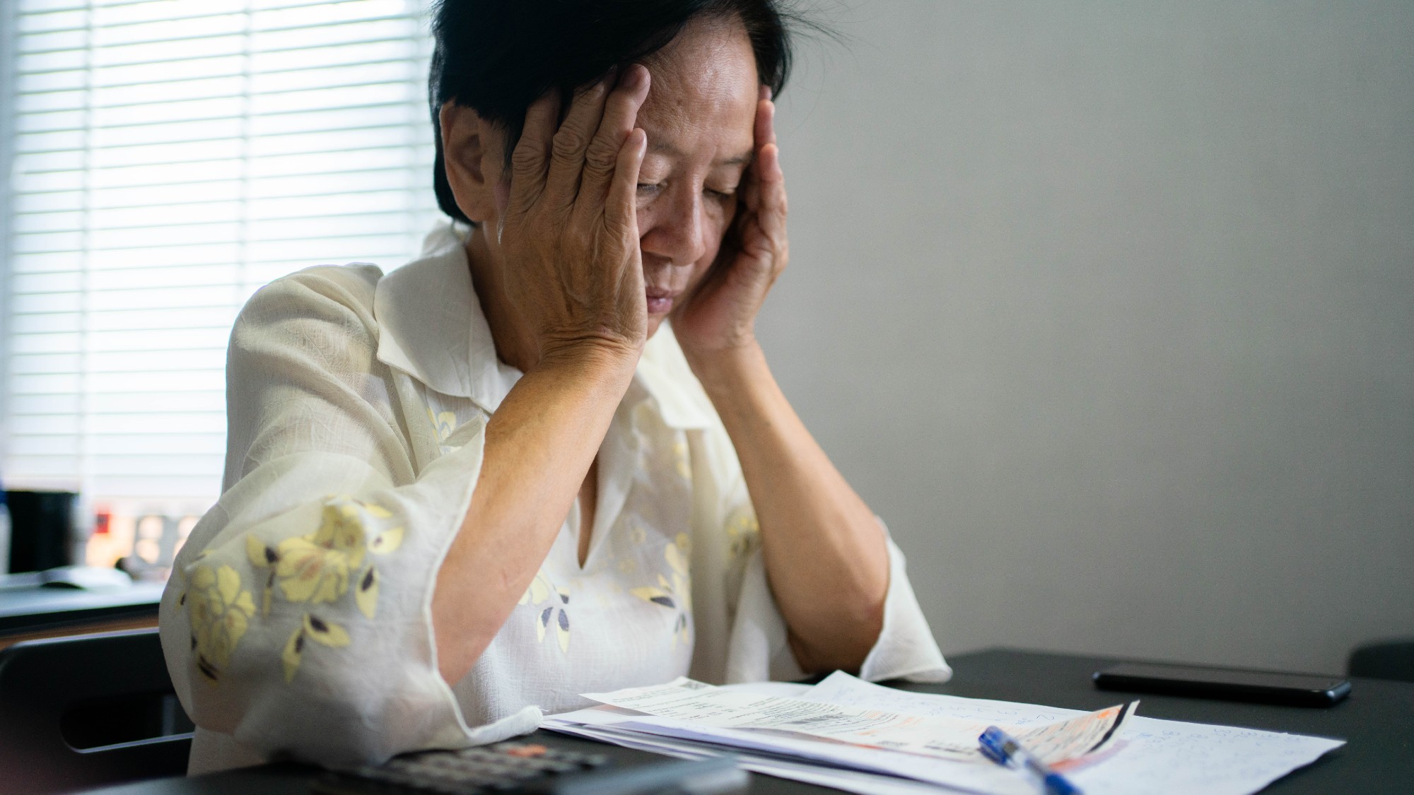 Senior woman sitting at living room table with her head in her hands, calculating expenses