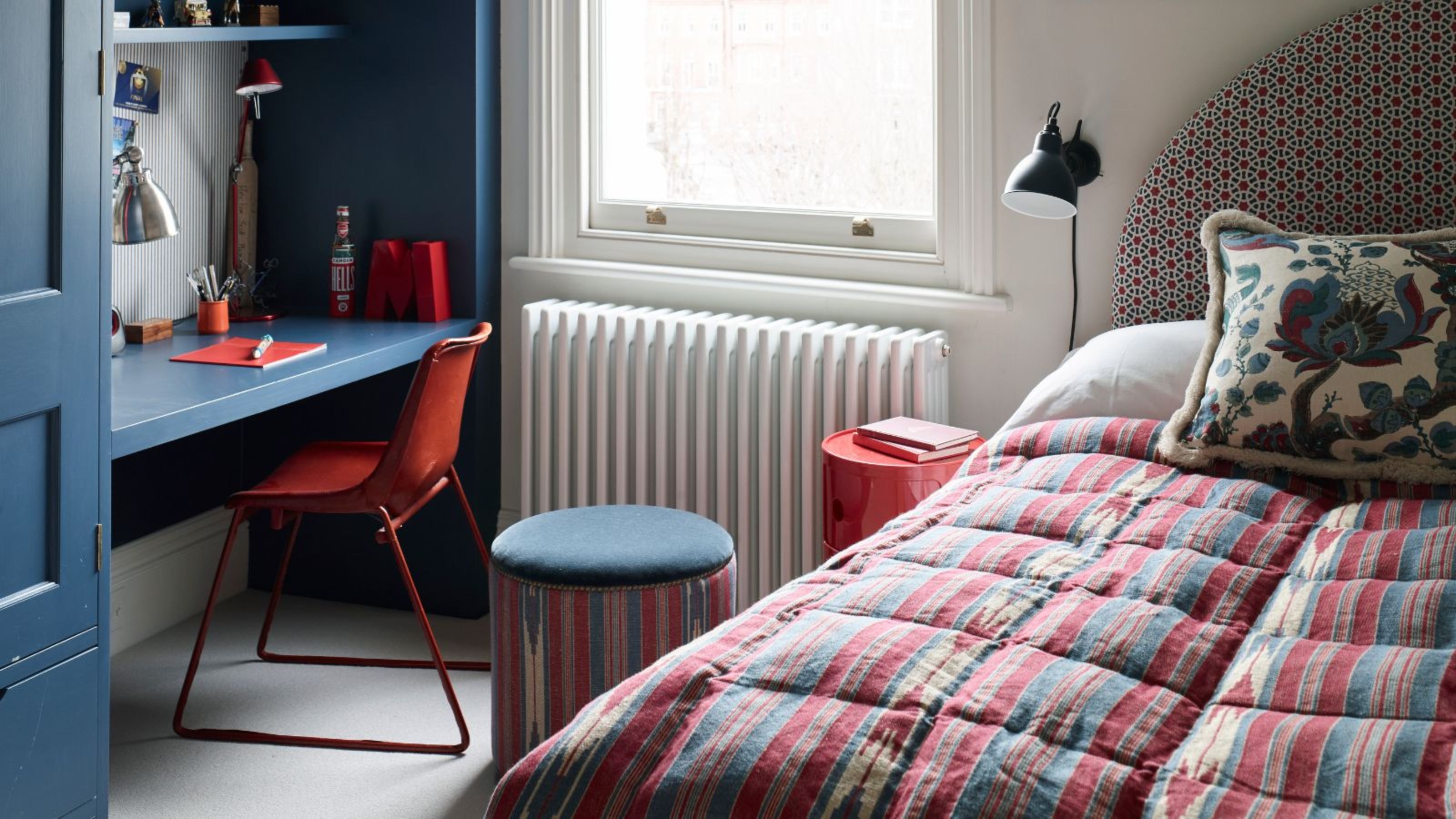 A small white bedroom with blue desk and closet, white radiator, large window, and patterned bedding on the single bed with a patterned cushion and curved patterned headboard.