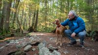 Man crouched down with his arm around Vizsla dog in the forest