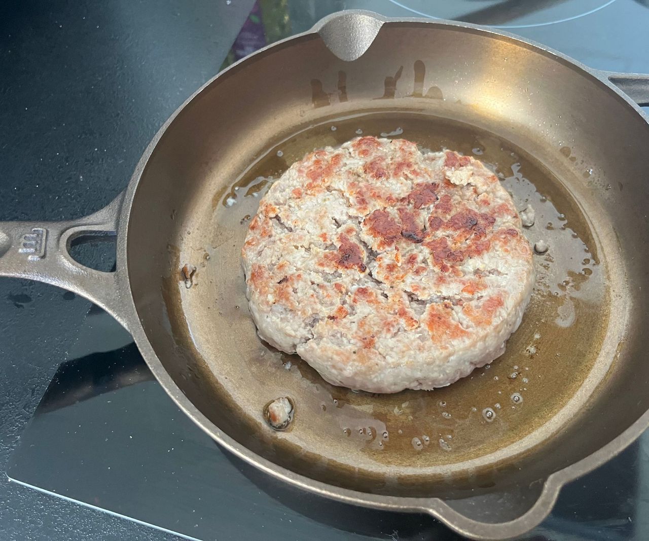 A small bronze colored cast-iron skillet on a black glass stove with a browned burger inside.