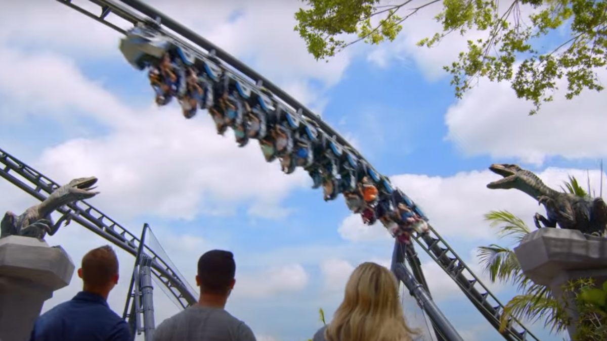 Guests walking under the track and in-between two Raptor statues at the Jurassic World VelociCoaster.