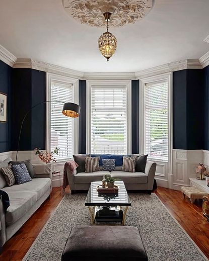 Blue living room with wooden floors neutral colored sofas and white bay windows with slatted white binds