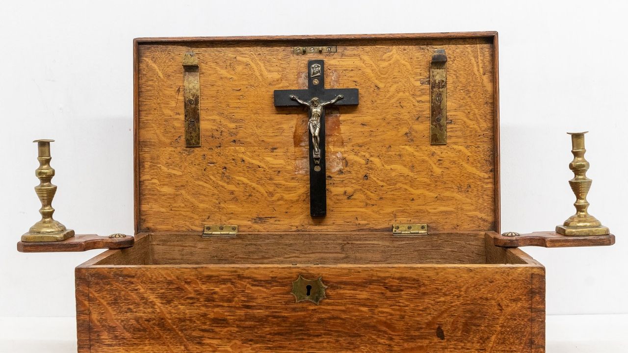 This is an open wooden chest. There is a black crucifix mounted on the underside of the lid. On each side there is a golden-looking candle holder mounted onto a wooden panel that swivels.