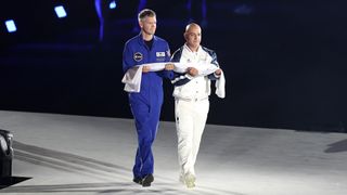 a man in a blue flight suit and a man in a white athletic tracksuit walk on a lighted stage carrying a folded flag