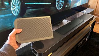 JBL Bar 1300X soundbar on TV stand and a hand holding a wireless surround speaker