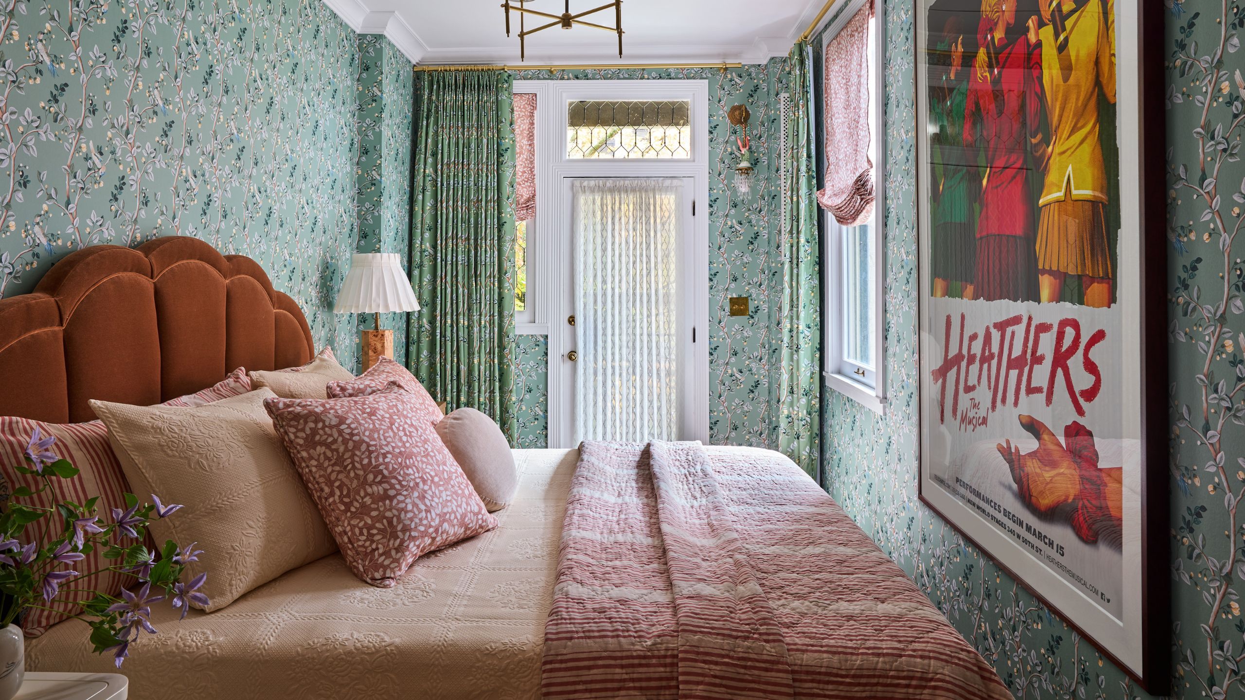 a large green floral wallpapered bedroom with rust colored scalloped bed, pink linens and a large the Heathers poster on the wall