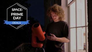 A man with long hair looking at a smartphone in between the Unistellar Odyssey Pro and a window at night, with a black Space.com Prime Day deals badge in the upper-left corner. 