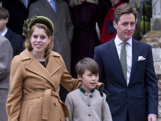 Princess Beatrice wearing a camel coat tied above her baby bump and a green headband, placing an arm around her stepson Christopher, who is standing next to dad Edoardo Mapelli Mozzi