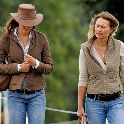 Kate Middleton wearing brown jacket and cowboy hat and walking next to Carole Middleton wearing a tan vest in front of green trees 