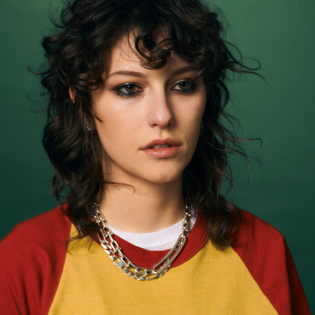 king princess poses with heavy eye makeup and wearing a yellow and red top with a chain necklace