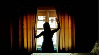 The image shows the back of a woman silhouetted against the bedroom curtains she is closing