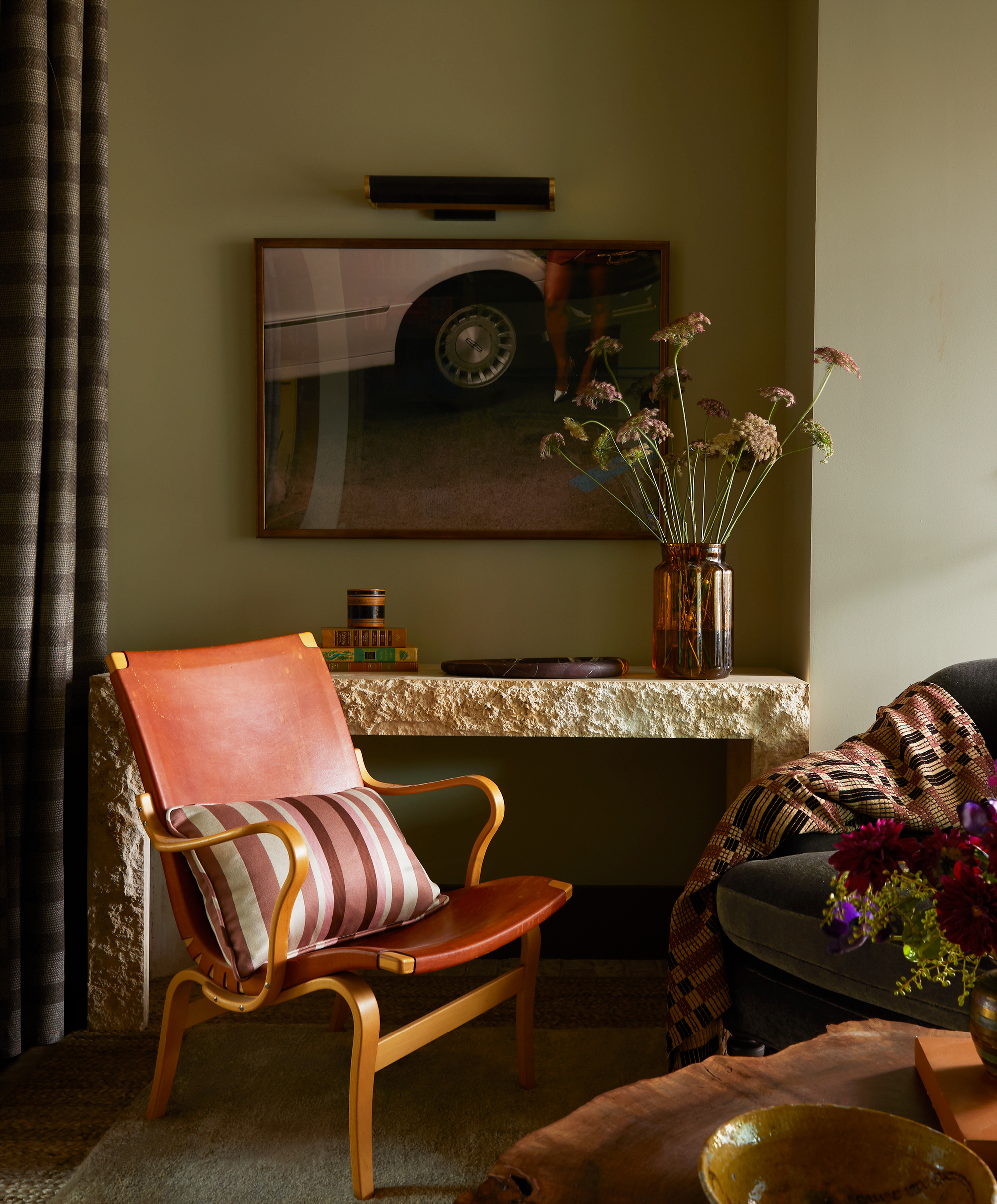 Image of the corner of a living room with sage green walls and a stone entryway table. There is a red leather accent chair with a striped throw pillow and a gray accent chair in view that has a patchwork quilt draped over the arm.