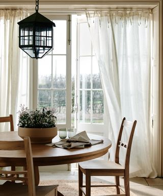 round wood dining room table with lantern pendant above and sheer linen curtains at window