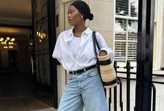 A woman looking to the side wearing a black headband, gold earrings, white tank top, white button-down shirt, black belt, and jeans and holding a raffia bag