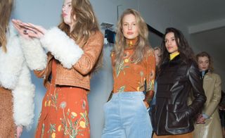 Models waiting in line for a fashion show to start, wearing bright orange shirts and dresses with wild dandelion prints, pastel blue corduroy pants, and black leather jackets from the Unique A/W 2015 collection.