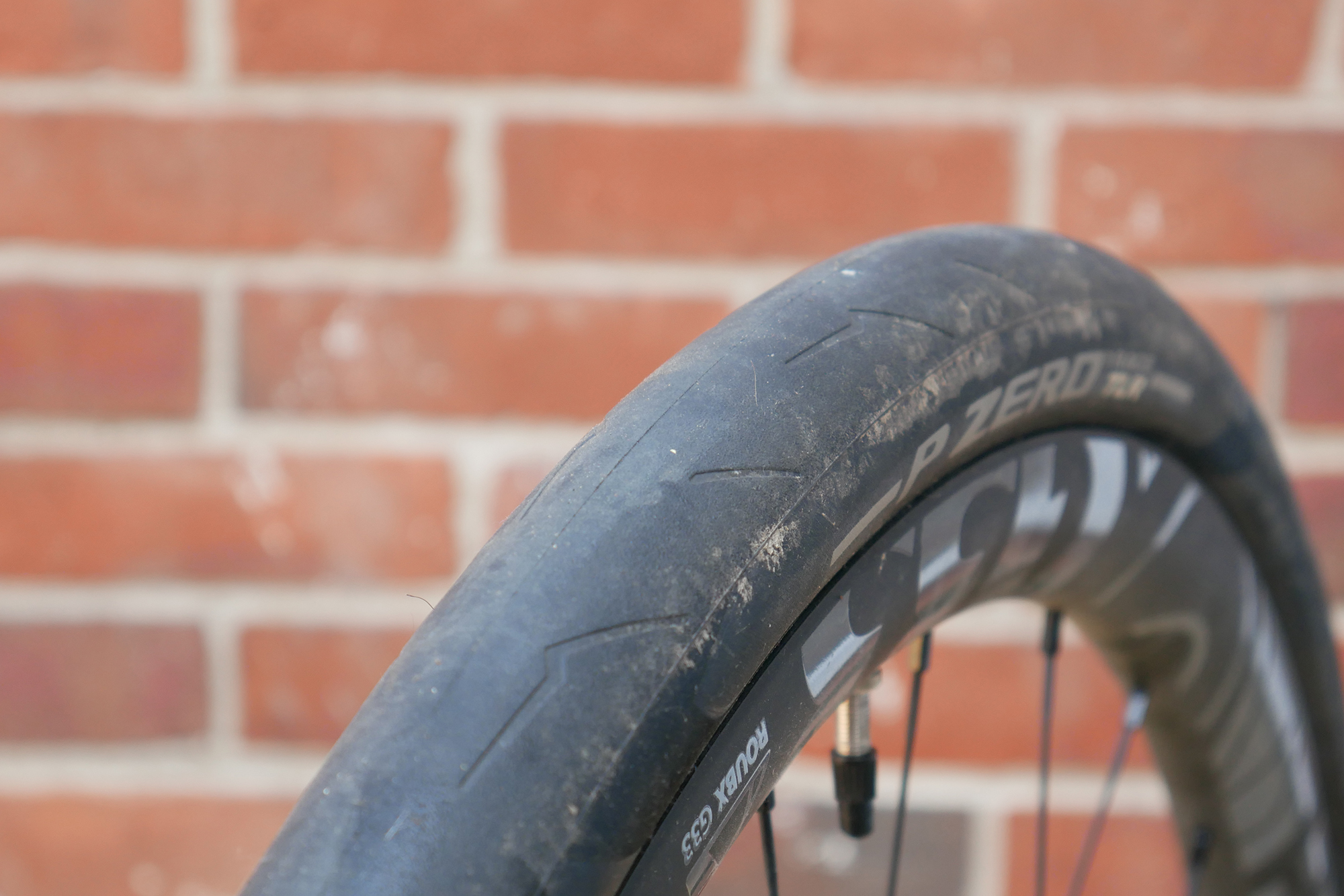 A black Pirelli P-Zero Race TLR tyre with white writing mounted to a Token wheel in front of a red brick wall showing the tread of the tyre