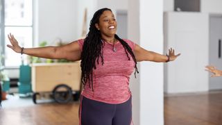 A woman in a pink shirt performs arm circles. She is smiling and has her arms reached out to the sides of her body and slightly behind her center.