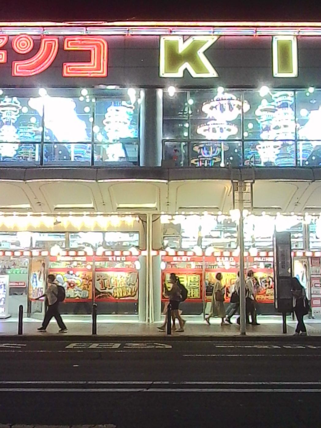 Brightly lit Japanese pachinko parlor at night with pedestrians walking past.