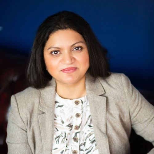 An image of Dr Hana Patel smiling at the camera, wearing a beige blazer and a floral shirt