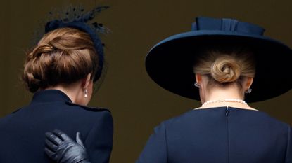 The back of Kate Middleton and Duchess Sophie wearing black coats and black hats as Sophie puts a gloved hand on Kate&#039;s back