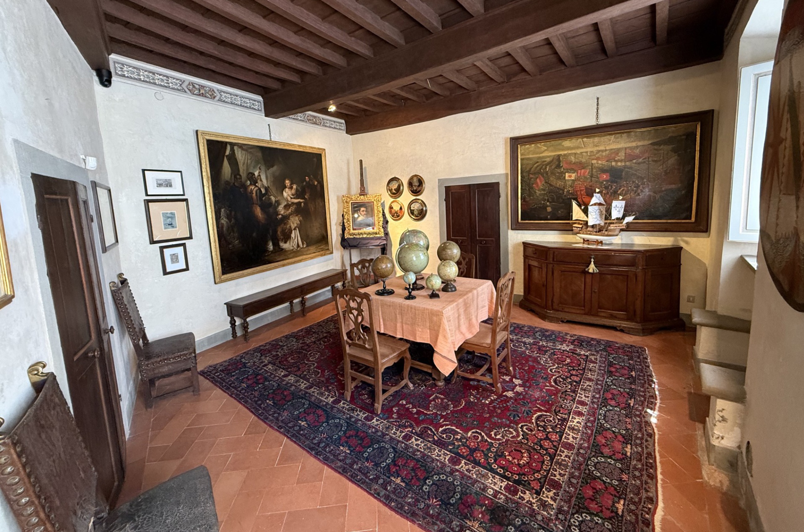 Museum room with a rug and a table with lots of globes on it, and old-looking pictures on the wall